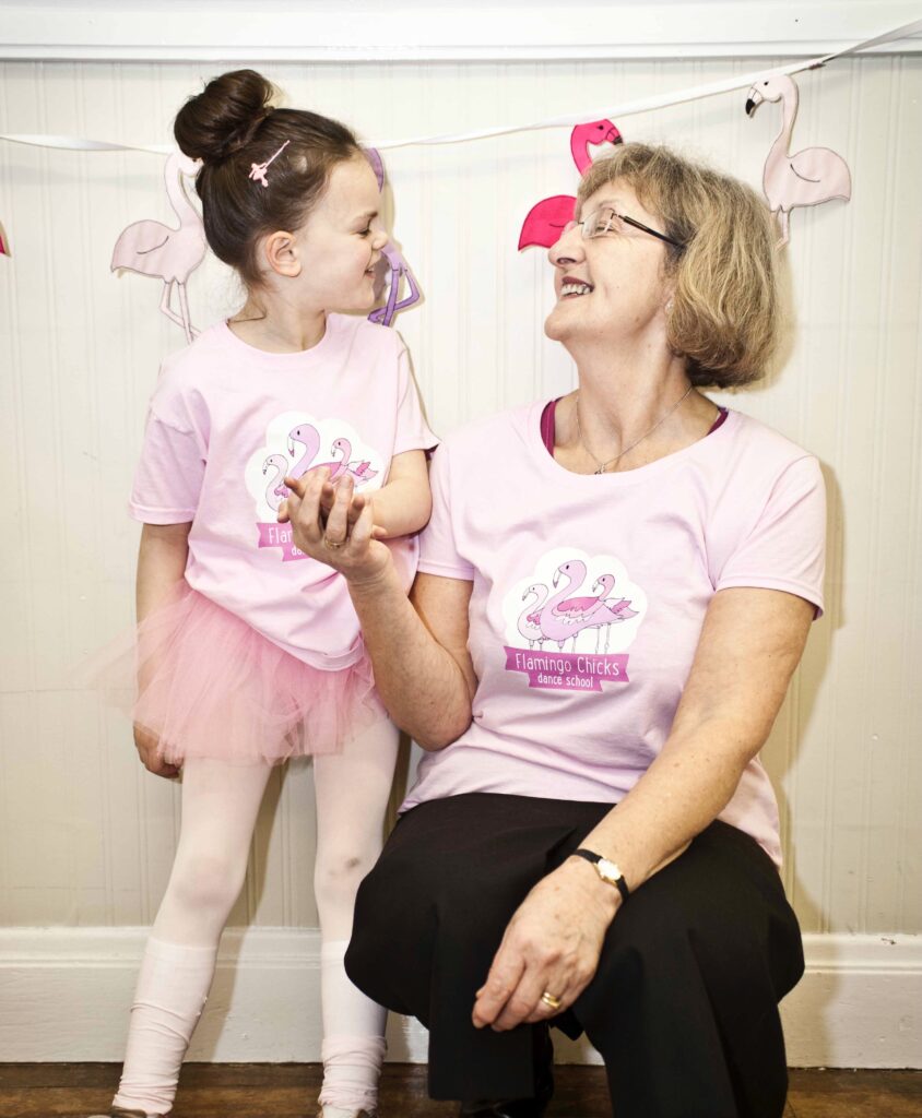 An older woman crouches next to a young girl in a ballet tutu and Flamingo Chicks t-shirt