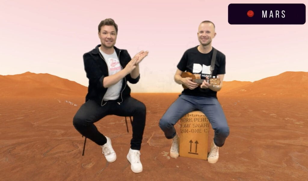 Two men are seated as they dance and sign in our virtual class. One is signing the letter 'M' for Mars and the the other is playing a guitar. A background of the planet mars is behind them. In the top right hand corner is the word 'Mars'.