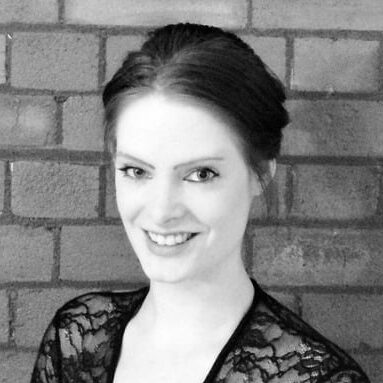 A black and white headshot of Lauren. She is fair skinned, has dark hair tied up, is wearing a black v-neck top, and is smiling.